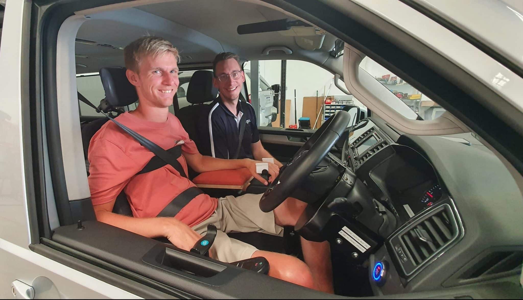 Two men in car smiling. The driver is wearing an orange t-shirt and is driving from wheelchair with hand controls.
